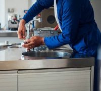 A shot of a man's hands working on a kitchen sink. He is wearing blue overalls with his tool kit in the background.